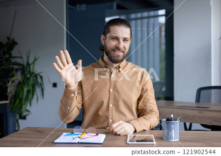 Smiling man in a casual shirt sitting at a desk in an office setting, waving at the camera, likely engaged in a friendly video conference or virtual meeting session. 121902544