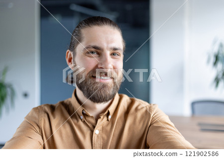 Happy young man with a beard taking a selfie in a contemporary office setting. Showcasing a friendly attitude, casual style, and a professional interior environment. 121902586