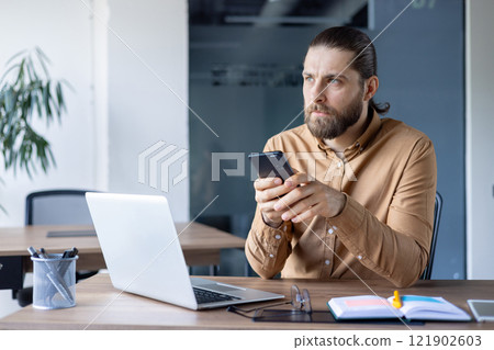 Focused man holds smartphone while working at an office desk with a laptop and documents. The office environment suggests analyzing, planning, and staying connected within a workplace. Focused man holds smartphone while working at an office desk with a laptop and documents. The office environment suggests analyzing, planning, and staying connected within a workplace. 121902603