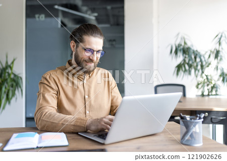 A male professional works confidently on a laptop at a wooden desk in a stylish office, focusing on business tasks with dedication. A male professional works confidently on a laptop at a wooden desk in a stylish office, focusing on business tasks with dedication. 121902626