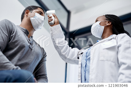 Female doctor consulting a male covid patient, taking his temperature with infrared thermometer in hospital room or clinic. Man wearing mask and health care professional pointing medical equipment. 121903046