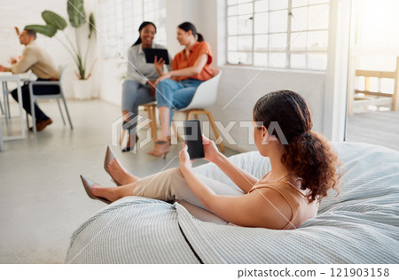 Relaxed, trendy and comfortable designer scrolling, texting and browsing on a phone while sitting on beanbag chair in a creative startup agency. Business woman on a break reading social media online 121903158