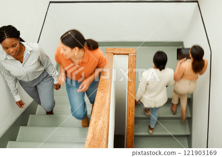 Overhead of busy, diverse female colleagues returning to work after break, with blurred digital tablet screen. Active business woman in motion talking and walking up office building stairs. 121903169