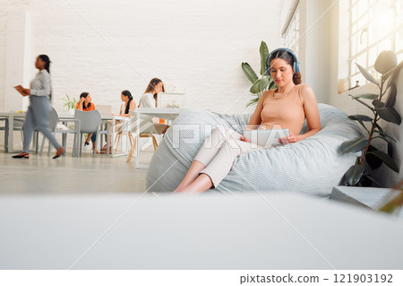 Creative intern browsing online with a tablet, listening to music on wireless headphones. Female on a break sitting on bean bags in a casual office space to improve creativity, work and mindfulness Creative intern browsing online with a tablet, listening to music on wireless headphones. Female on a break sitting on bean bags in a casual office space to improve creativity, work and mindfulness 121903192