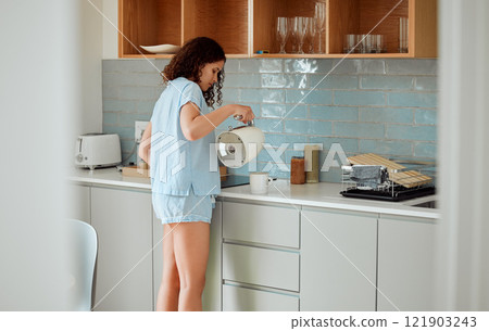 Young woman having coffee, tea or morning routine in comfortable pajamas in her stylish kitchen at home. Millennial girl starting her relaxing day with caffeine or beverage in her apartment 121903243