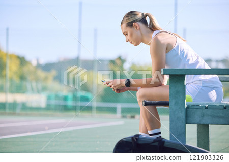 Fit tennis player with phone checking fitness goal progress on a sports exercise app online while taking a break at the court. A sportswoman checking messages on cellphone and waiting for a coach Fit tennis player with phone checking fitness goal progress on a sports exercise app online while taking a break at the court. A sportswoman checking messages on cellphone and waiting for a coach 121903326