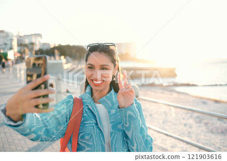 Smiling, trendy and fun woman, tourist or influencer taking selfie with peace sign gesture on phone for social media, video call or photo. Exploring, traveling and sightseeing at promenade outdoors 121903616