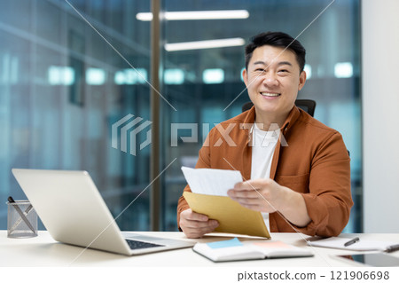 Portrait of Asian man inside office at workplace, man looking at camera received joyful news message. Businessman holding mail envelope letter with notification in hands. Portrait of Asian man inside office at workplace, man looking at camera received joyful news message. Businessman holding mail envelope letter with notification in hands. 121906698