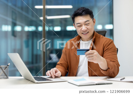 Man at workplace inside office with laptop and bank credit card. Asian man buys online and books services remotely. Businessman smiling happily. 121906699