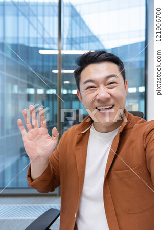 Video call, Asian man looking into phone camera and talking to colleagues using smartphone app. Businessman at workplace inside office. 121906700