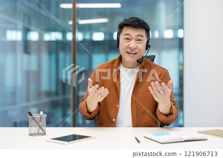 Webcam view, man with headset looking at camera, talking to colleagues and partners remotely. Asian businessman smiling at online business meeting conference. 121906713