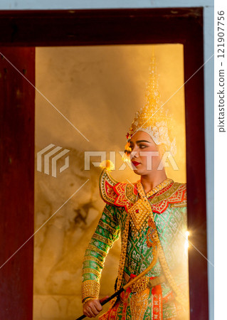 Vertical image of man with Khon mask charactor as deity stand with look to left side in front of door frame of old building with beam warm light in the background. 121907756