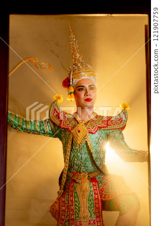 Vertical image of man with Khon mask charactor as deity stand with performance action in front of door frame of old building with beam warm light in the background. 121907759