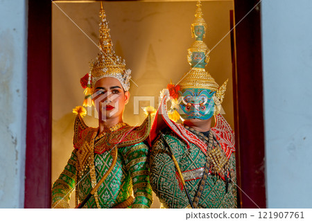 Two charactors of Khon mask costume stand with backs leaning against each other and giant mask stand in right side at door frame with beam light in the back. 121907761