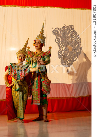 Vertical image of man and woman with Khon or Mask dance costume stand with performance action in front of grand shadow play in the background with white screen. Vertical image of man and woman with Khon or Mask dance costume stand with performance action in front of grand shadow play in the background with white screen. 121907802