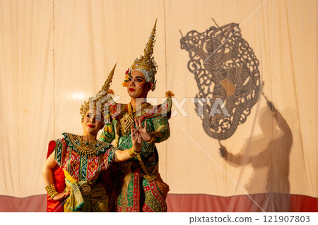 Man and woman with Khon or Mask dance costume stand with performance action in front of grand shadow play in the background with white screen. Man and woman with Khon or Mask dance costume stand with performance action in front of grand shadow play in the background with white screen. 121907803