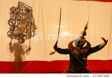 Man with Khon or Mask dance stand with performance action in front of grand shadow play in the background with white screen. Man with Khon or Mask dance stand with performance action in front of grand shadow play in the background with white screen. 121907806