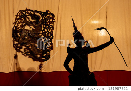 Close up and silhouette of Khon or Mask dance player stand with performance action in front of grand shadow play in the background with white screen. Close up and silhouette of Khon or Mask dance player stand with performance action in front of grand shadow play in the background with white screen. 121907821