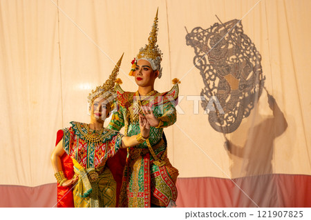 Close up man and woman with Khon or Mask dance costume stand with performance action in front of grand shadow play in the background with white screen. Close up man and woman with Khon or Mask dance costume stand with performance action in front of grand shadow play in the background with white screen. 121907825
