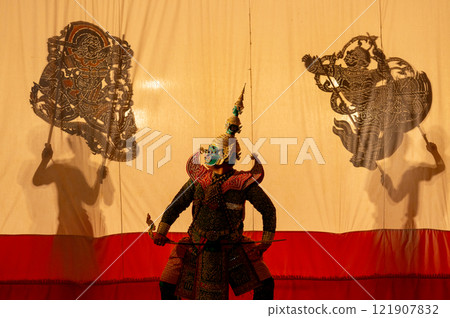 Low light of man as giant charactor with Khon or Mask dance costume stand with performance action in front of two set of grand shadow play in the background with white screen. Low light of man as giant charactor with Khon or Mask dance costume stand with performance action in front of two set of grand shadow play in the background with white screen. 121907832