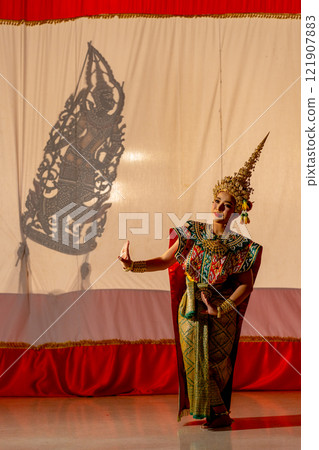 Vertical image of one woman with Khon or Mask dance dress stand with performance action in front of grand shadow play in the background with white screen. Vertical image of one woman with Khon or Mask dance dress stand with performance action in front of grand shadow play in the background with white screen. 121907883