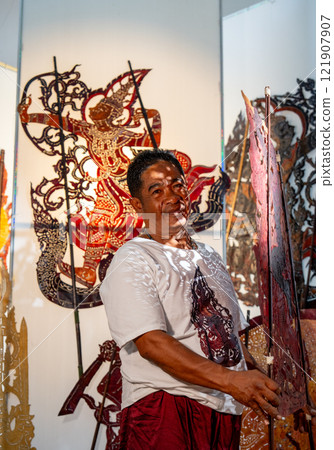 Vertical image of Asian senior man hold the piece of grand shadow play craft and look at camera also stay in area of workplace and he look pround for his work. 121907907