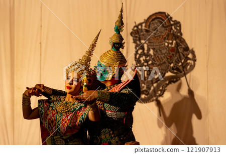 Man as giant charactor and woman with Khon or Mask dance costume stand with performance action in front of grand shadow play in the background with white screen. 121907913