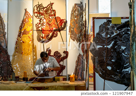 Asian senior man use hammer and chisel to create grand shadow play craft and he concentrate to work in area of workplace with several products are place around workplace area. 121907947