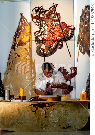 Vertical image of Asian senior man use hammer and chisel to create grand shadow play craft and he concentrate to work in area of workplace with several products are place around workplace area. 121907948