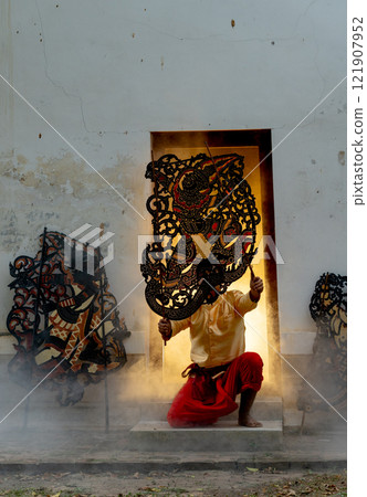 Vertical image of one man hold craft of grand shodow play charactor and sit in front of  door frame also some crafts in front of old building and warm light in the back of room. 121907952