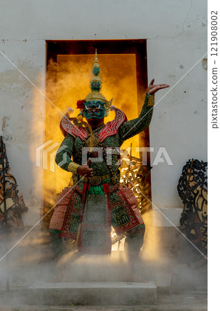 Vertical image of one man with Khon mask charactor giant stand and performance action in front of door frame of old building also some crafts in front of old building and warm light in the back 121908002