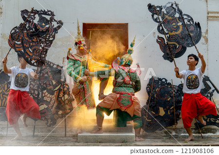 Two men with Khon mask charactor as angel and giant stand and performance action as figthing in front of door frame of old building also two men hold grand shadow play in front and warm light in back. 121908106