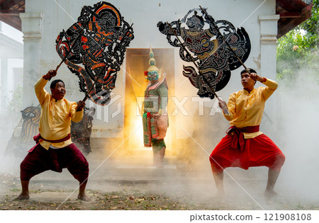 One man with Khon mask charactor as giant stand in front of door frame of old building also two men hold grand shadow play in front and warm light in back. 121908108
