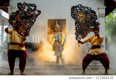 One man with Khon mask charactor as angel stand in front of door frame of old building also two men hold grand shadow play in front and warm light in back. 121908110