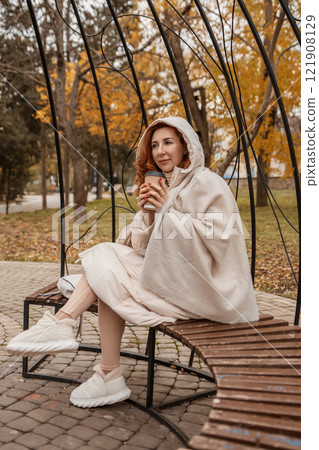 Woman Autumn Park Beige Poncho: Enjoying Coffee Outdoors Fall Day Relaxing Cozy Outfit. Woman Autumn Park Beige Poncho: Enjoying Coffee Outdoors Fall Day Relaxing Cozy Outfit. 121908129