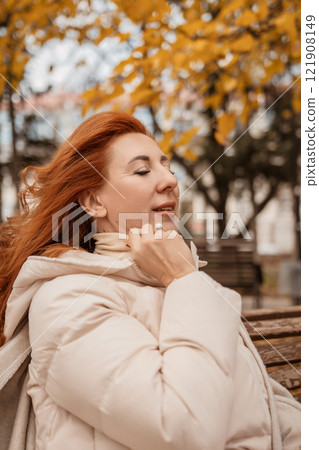Woman Autumn Park Relaxation: Redhead enjoys fall breeze, sitting bench, serene moment. Woman Autumn Park Relaxation: Redhead enjoys fall breeze, sitting bench, serene moment. 121908149