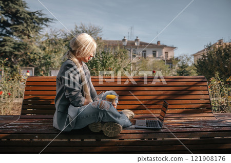A woman sits on a bench with a laptop open in front of her. She has a cup of coffee in her hand and her jeans are ripped. The scene suggests a relaxed and casual atmosphere. 121908176