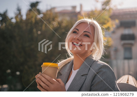 A blonde woman sits on a bench drinking coffee from a yellow cup. She is wearing a gray jacket and has her hair in a ponytail. The scene is peaceful and relaxing. A blonde woman sits on a bench drinking coffee from a yellow cup. She is wearing a gray jacket and has her hair in a ponytail. The scene is peaceful and relaxing. 121908279