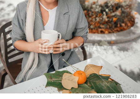 A woman is sitting at a table with a cup of coffee and a plate of oranges. The oranges are on a leaf, and the woman is wearing a gray jacket. Concept of relaxation and enjoyment. 121908283
