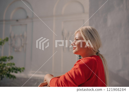 A woman in a red sweater is sitting on a ledge. She is smiling and looking out at the street. 121908284