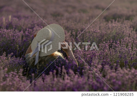 A woman is sitting in a field of purple flowers, wearing a straw hat A woman is sitting in a field of purple flowers, wearing a straw hat 121908285
