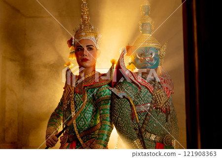 Wide shot of two charactors of Khon mask costume stand with backs leaning against each other and giant mask stand in right side at door frame with beam light in the back. 121908363