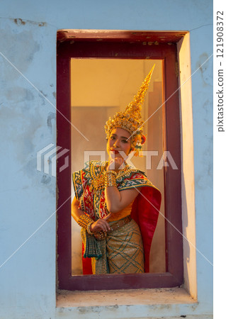 Vertical image of one woman with Khon Mask costume stand with shy action at window frame with warm light in the back. Vertical image of one woman with Khon Mask costume stand with shy action at window frame with warm light in the back. 121908372