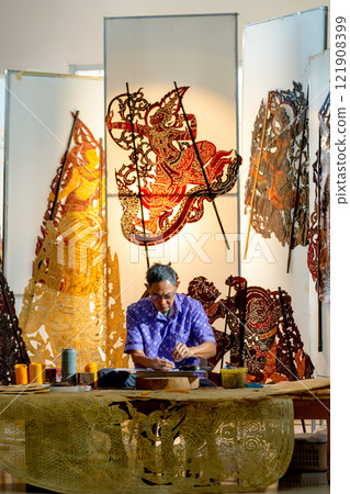 Vertical image Asian senior man wear glasses use hammer and chisel to create grand shadow play craft and he concentrate to work in area of workplace with several products are place around workplace. 121908399