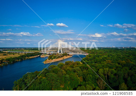 Cable stayed bridge over river surrounded by lush green forest. Redzinski bridge over Odra river with cars driving on highway in Wroclaw, Poland Cable stayed bridge over river surrounded by lush green forest. Redzinski bridge over Odra river with cars driving on highway in Wroclaw, Poland 121908585