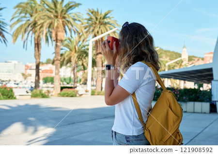 Female traveler in casual clothing taking picture in coastal town. Woman using orange retro instant camera to take photo during vacation. Tourist captures memories during travel Female traveler in casual clothing taking picture in coastal town. Woman using orange retro instant camera to take photo during vacation. Tourist captures memories during travel 121908592