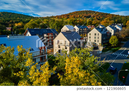 Residential building facade with balconies and windows near vibrant autumn forest, aerial view. Contemporary apartments in suburban eco friendly neighborhood with green spaces near mountains 121908605