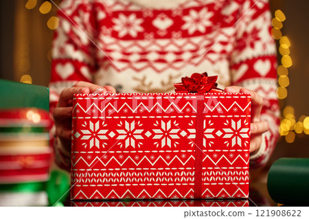 Woman wearing Christmas sweater holding wrapped Christmas gift with wrapping paper and ribbon table. Preparing presents for winter holidays 121908622