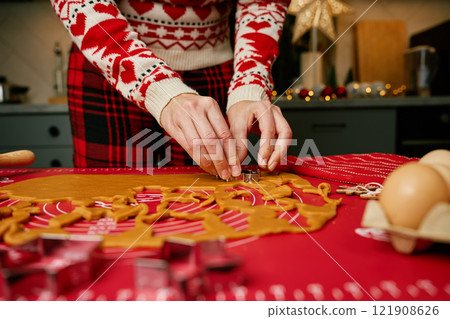 Woman in festive sweater using cookie cutters to shape gingerbread dough on red mat. Baking traditional Christmas cookies in home kitchen Woman in festive sweater using cookie cutters to shape gingerbread dough on red mat. Baking traditional Christmas cookies in home kitchen 121908626