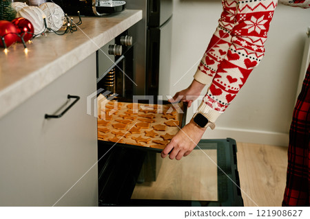 Woman in red festive sweater putting tray of Christmas cookies into oven. Process of cooking traditional gingerbread cookies for winter holidays at home kitchen Woman in red festive sweater putting tray of Christmas cookies into oven. Process of cooking traditional gingerbread cookies for winter holidays at home kitchen 121908627